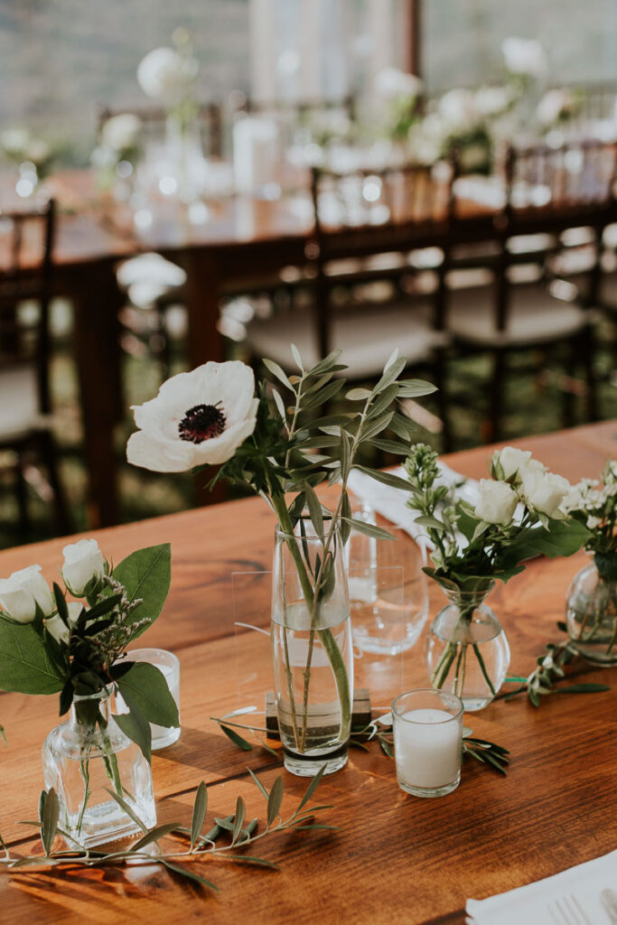 white poppy and sage leaves sit beside small white roses on a wooden reception table at a wedding