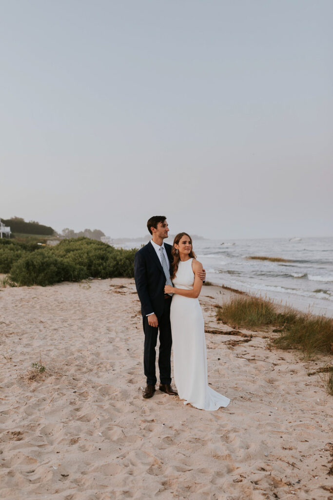 a Cape Cod groom looks out at the ocean on Cape Cod with his arm around his bride
