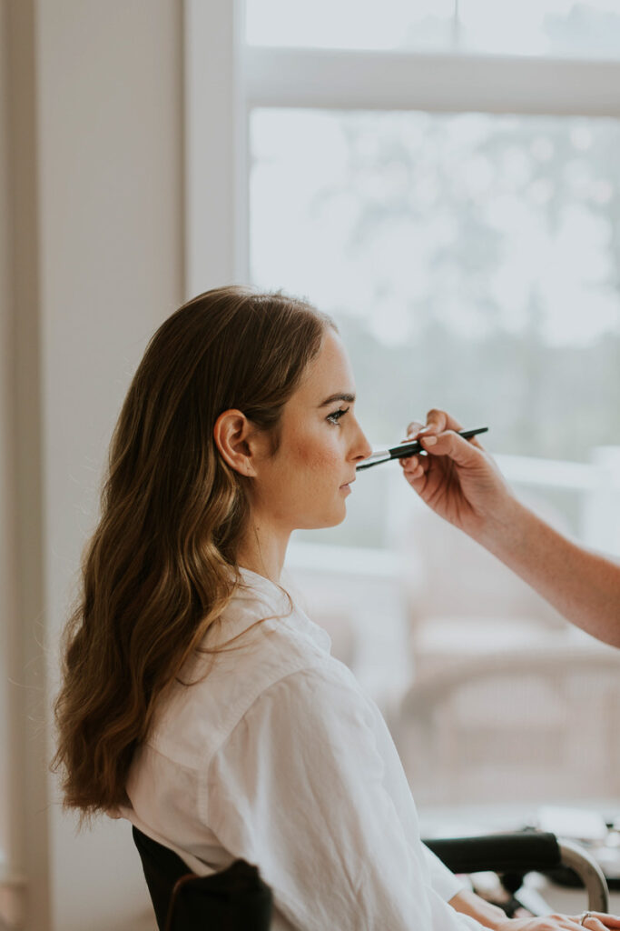 bride sits in chair as makeup is applied during her wedding day