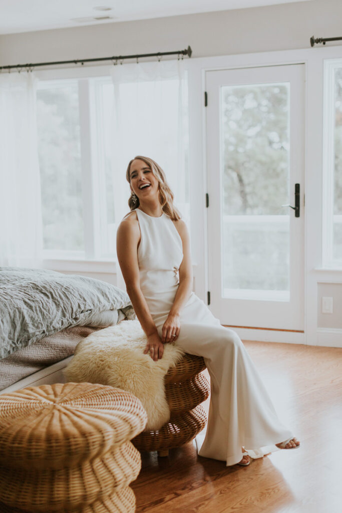 bride poses on wicker chair and laughs at something her bridesmaids are saying