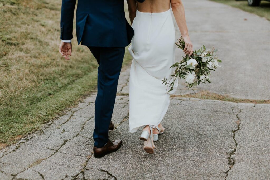 groom escorts bride down the driveway while she dangles her flowers from one hand