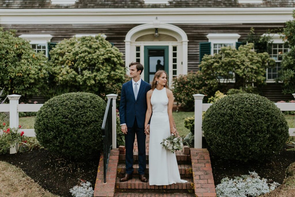 Bride and groom hold hands and look in opposite directions on steps of Cape Cod home