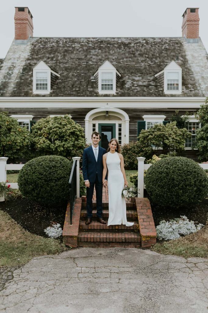 bride and groom pose holding hands in front of quaint Cape Cod cottage