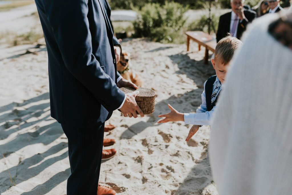 little boy ring bearer hands the basket with the rings to one of the Groomsmen during Cape Cod outdoor ceremony